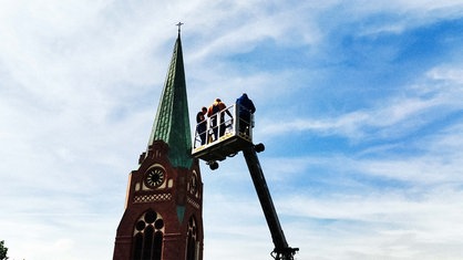 Ein Kirchturm, davor eine Krangondel, in der sich mehrere Menschen befinden