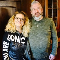 Eine blonde Frau mit Brille steht neben einem Mann mit kurzen, grauen Haaren in einer Kneipe.