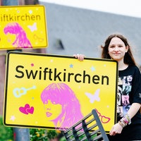Die Schülerin Aleshanee Westhoff zeigt das Ortsschild "Swiftkirchen" zu Ehren der Musikerin Taylor Swift auf dem Heinrich-König-Platz in Gelsenkirchen in die Kamera.
