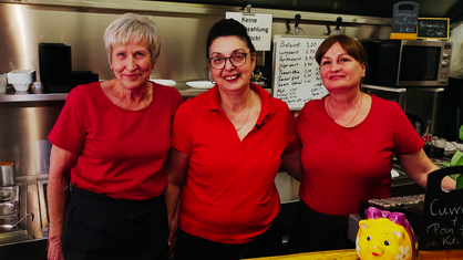 Die drei Frauen Moni, Suzi und Mira stehen in roten T-Shirts nebeneinander hinter dem Tresen des Imbisses und lächeln.