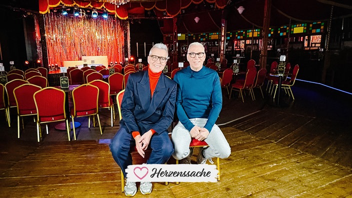 Knut Vanmarcke und Dirk Vossberg-Vanmarcke auf einem Stuhl in ihrem Theater. Im Hintergrund ist die Bühne erkennbar.