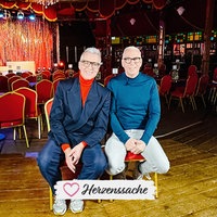 Knut Vanmarcke und Dirk Vossberg-Vanmarcke auf einem Stuhl in ihrem Theater. Im Hintergrund ist die Bühne erkennbar.
