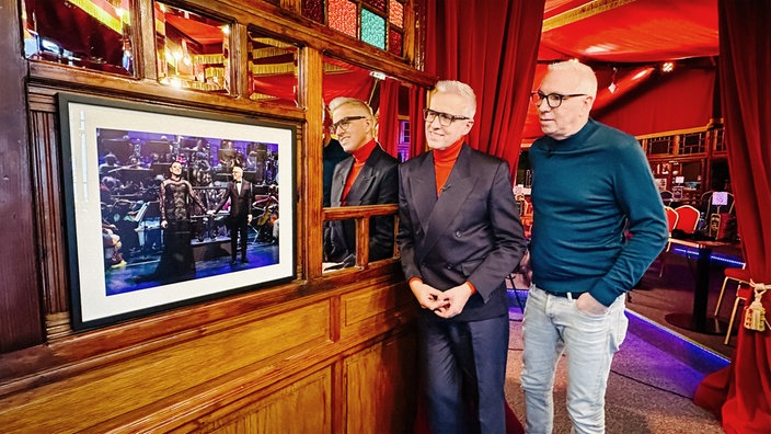 Knut Vanmarcke und Dirk Vossberg-Vanmarcke blicken auf ein eingerahmtes Bild ihres größten Auftritts. Im Hintergrund ihr Theater.