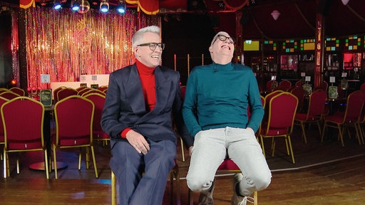 Knut Vanmarcke und Dirk Vossberg-Vanmarcke sitzen auf zwei Stühlen in ihrem Theater in Bonn.