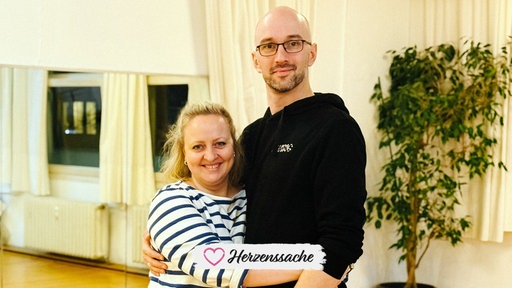 Judith Buschfeld, eine blondhaarige Frau im schwarz-weiß gestreiften Pullover, und László Tarkanyi, ein großer Mann mit Dreitagebart, Brille und schwarzem Kapuzenpullover, stehen nebeneinander und umarmen sich.