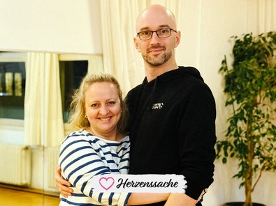 Judith Buschfeld, eine blondhaarige Frau im schwarz-weiß gestreiften Pullover, und László Tarkanyi, ein großer Mann mit Dreitagebart, Brille und schwarzem Kapuzenpullover, stehen nebeneinander und umarmen sich.