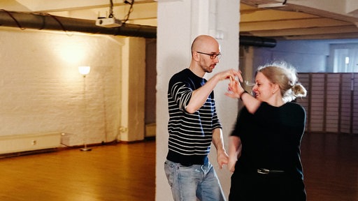 Judith Buschfeld, eine blondhaarige Frau im schwarzen Pullover, und László Tarkanyi, ein großer Mann mit Dreitagebart, Brille und gestreiftem Pullover tanzen gemeinsam.