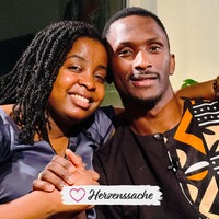 Michèle Leutcha Tchatchou, eine junge Frau in einer violetten Bluse und Archimede Mbazou Kouadjio, ein junger Mann in einem braunen gemusterten Hemd, sitzen innig umarmt nebeneinander und halten sich die Hände.