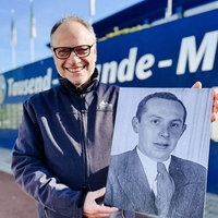 Alfred Brechner steht vor der Tausend Freunde Mauer am Schalker Stadion. Er hält ein Foto von Ernst Alexander hoch und lächelt.