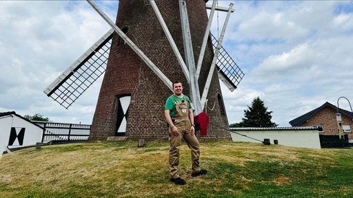 Der Müller Leon Joachims steht in brauner Arbeitskleidung vor seiner historischen Windmühle.