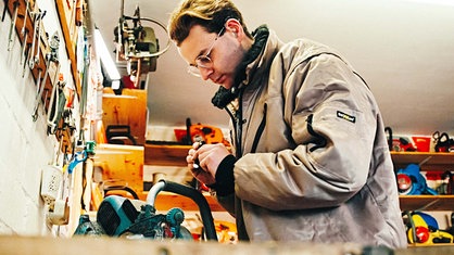 Luis Hantschel, ein junger, braunhaariger Mann in einer beigen Jacke, beugt sich mit einer Taschenlampe über eine Werkbank, auf der eine türkisfarbene Kettensäge liegt.