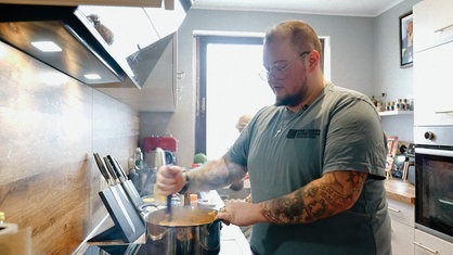 Melvin Flor steht in der Küche vor einem Topf auf dem Herd und rührt in ihm.