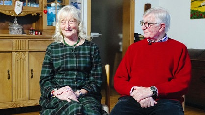 Heidi und Horst Thielke, ein älteres Ehepaar mit silbernen Haaren, sitzt im Wohnzimmer nebeneinander. Heidi spricht und ihr Mann Horst schaut sie freundlich von rechts an.