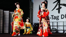 Eine Gruppe aus drei japanischen Frauen tanzt im Kimono und mit Fächern auf einer Bühne.