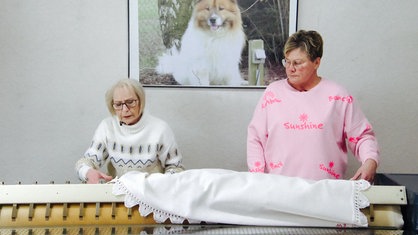 Zwei Frauen stehen nebeneinander, vor ihnen liegt ein großes weißes Laken auf einem Tisch.