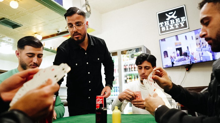 Jendi Mahmud steht an einem Tisch, an dem mehrere Männer sitzen. Sie halten Karten in der Hand.