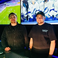 Roman und Kirill hinter der Theke der Youbar. Hinter ihnen hängen große Bildschrime an den Wänden, die Sport zeigen. 
