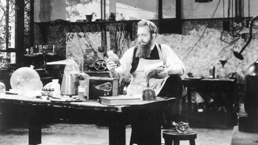 Aufnahme von Wilhelm Conrad Roentgen in seinem Labor. Er hält ein Buch in der linken und schaut eine Glasflasche in der rechten Hand an.
