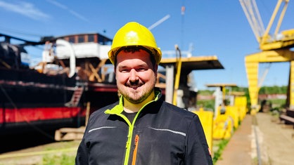 Sebastian Winter in der Halbtotalen. Er trägt einen Sicherheitshelm und Arbeitskleidung. Im Hintergrund wird das Schiff "Oscar Huber" restauriert.