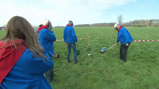 Einwohner von Herzebrock-Clarholz stehen mit Ohrenschützern auf einem freien Feld, einer hält eine brennende Fackel an den Boden einer Milchkanne.