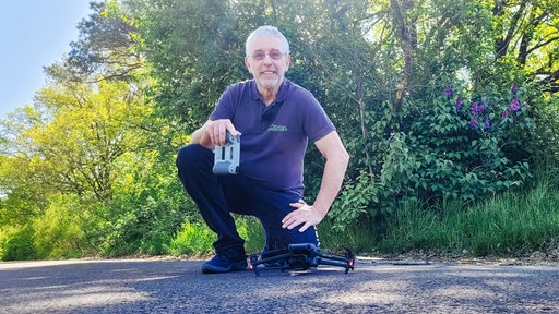 Dietmar Meier kniet im Grünen und lächelt in die Kamera. Vor ihm liegt seine Drohne in der Hand hält er die Fernsteuerung.