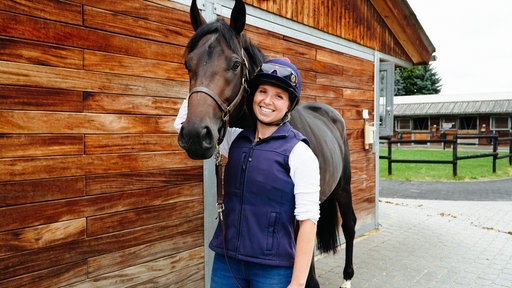 Chantal Warmer trägt Reitkleidung und steht neben einem dunkelbraunen Pferd vor einem Stall.