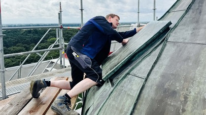 Dachdecker Gerald Plenter arbeitet an den Dachziegeln des Raesfelder Schlosses. Er schaut konzentriert auf die vor ihm befestigten Ziegel. Im Hintergrund sieht man einen Teil des Gerüsts und die umliegende Landschaft.