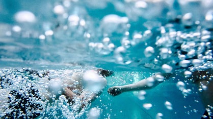 Harald Peters schwimmt unter Wasser. Neben ihm steht eine weitere Person, die unterstützend ihren Arm ausstreckt.