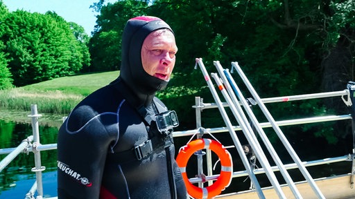 Ein Mann in schwarzem Neoprenanzug, an dem eine kleine Kamera befestigt ist. Im Hintergrund sieht man Gerüststäbe an ein Gerüst lehnen, an dem ein Rettungsring hängt, dahinter blickt man auf Wasser und Grünflächen.