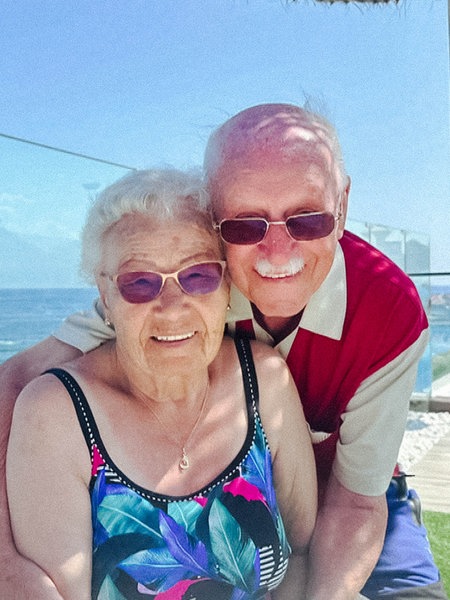 Regina Schmer, eine ältere Frau mit weißen Locken mit Sonnenbrille und Badeanzug, sitzt. Hinter ihr steht Rudi Vollak, ein älterer Mann mit Sonnenbrille, und umarmt die Frau. 