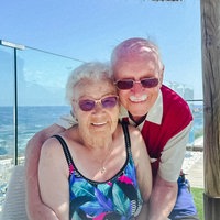 Regina Schmer, eine ältere Frau mit weißen Locken mit Sonnenbrille und Badeanzug, sitzt. Hinter ihr steht Rudi Vollak, ein älterer Mann mit Sonnenbrille, und umarmt die Frau. 
