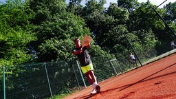 Ein Mann steht in Sportklamotten auf einem Sportplatz und hält einen Holzschläger und einen Ball vor sich.