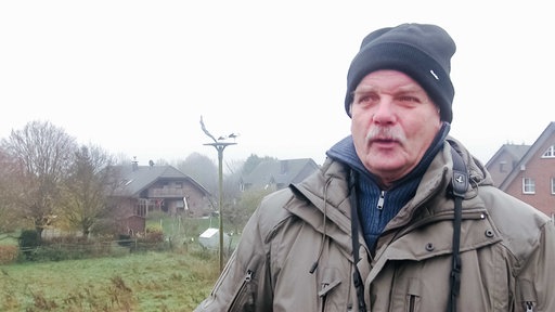 Mann blickt nach vorne, er spricht. Im Hintergrund links sieht man verschwommen ein Strochennest