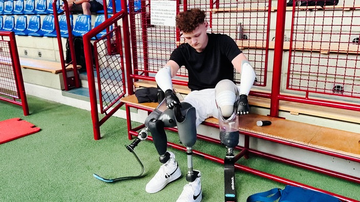 Fabian Abels sitzt auf einer Bank in einer Sporthalle und zieht seine Beinprothesen an