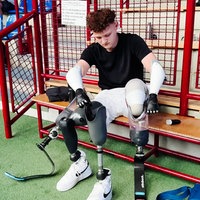 Fabian Abels sitzt auf einer Bank in einer Sporthalle und zieht seine Beinprothesen an