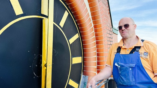Ein Mann in blauer Latzhose steht neben dem Zifferblatt einer Kirchturmuhr