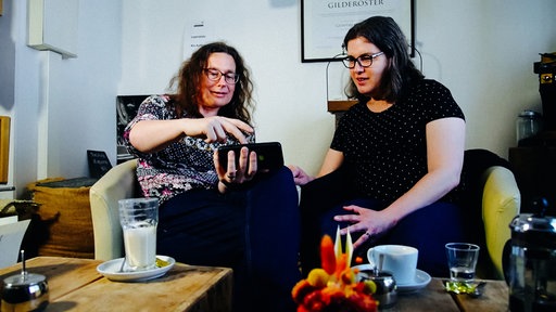 Andrea und Claire Stark sitzen in Sesseln und schauen Bilder auf einem Handy an, das Andrea in der Hand hält.