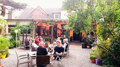 Auf dem Foto sieht man den Biergarten der Kastanie am Hax. Kneipengäste sitzen an Tischen umgeben von Bäumen.