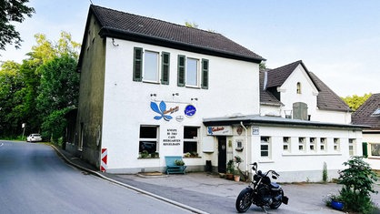Zu sehen ist die Kneipe ,,Kastanie am Hax". Es ist ein zweistöckiges Haus mit dem Eingangsbereich an der Seite. Die Kneipe steht direkt an einer Straße.