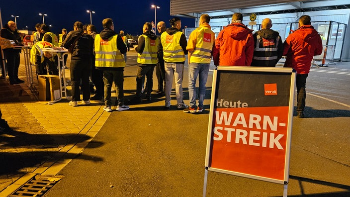 Demonstrantinnen und Demonstranten stehen vor dem Flughafen. Davor ein Schild mit der Aufschrift "verdi - Warnstreik - Heute"