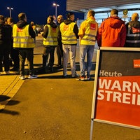 Demonstrantinnen und Demonstranten stehen vor dem Flughafen. Davor ein Schild mit der Aufschrift "verdi - Warnstreik - Heute"