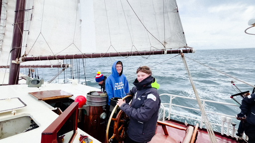 Malte Heidbrink in einer dunkelblauen Regenjacke steht am Steuer des Segelboots Regina Maris, hinter ihm zwei weitere Schüler