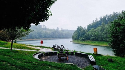 An einer Talsperre regnet es. Im Vordergrund ist ein Kreis aus Bänken. Dahinter ist der See.