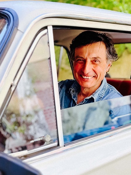 Andrea Giacin sitzt auf dem Fahrsersitz seinen umgebauten Ladas. Die Fensterscheibe ist heruntergelassen, er hält das Lenkrad und lächelt zufrieden in die Kamera. 