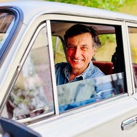 Andrea Giacin sitzt auf dem Fahrsersitz seinen umgebauten Ladas. Die Fensterscheibe ist heruntergelassen, er hält das Lenkrad und lächelt zufrieden in die Kamera. 