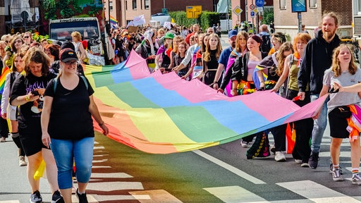 Eine Menschenmenge, die eine Straße entlangläuft. In der Mitte halten einige Personen eine große Regenbogenflagge.