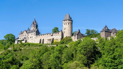Blick auf die Burgfassade der Burg Altena, die umgeben ist von grünen Büschen und Bäumen am Hang, Burg selbst mit Mauer und zwei hohen Türmen