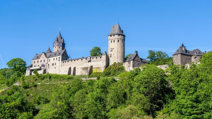 Blick auf die Burgfassade der Burg Altena, die umgeben ist von grünen Büschen und Bäumen am Hang, Burg selbst mit Mauer und zwei hohen Türmen