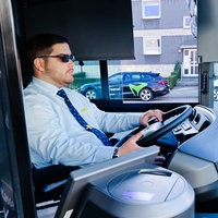 Kilian Zander, hellblaues Hemd, blaue Krawatte, schwarze Sonnenbrille, sitzt am Steuer eines Busses und blickt durch die Frontscheibe, im Hintergrund parken Autos auf der Straße