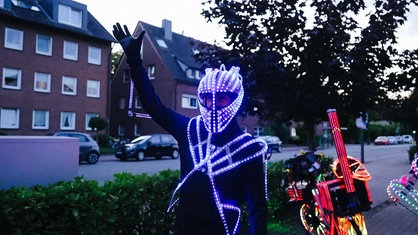 Ein Mann mit einem Motorradhelm hebt die Hand in die Luft. Er ist mit LED- Lichtern geschmückt.